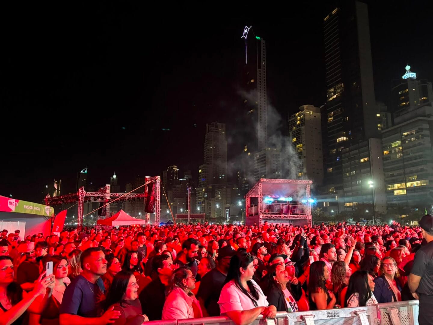 Vis&atilde;o de dentro da plateia lotada durante o show &agrave; noite, com luzes de palco sobre a multid&atilde;o, palco iluminado com Alexandre Pires ao longe e pr&eacute;dios da cidade ao fundo.