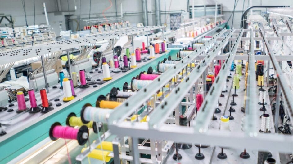 Linha de produção industrial de fábrica têxtil com carretéis de fios coloridos instalados em máquinas de tear e bordado automatizado.