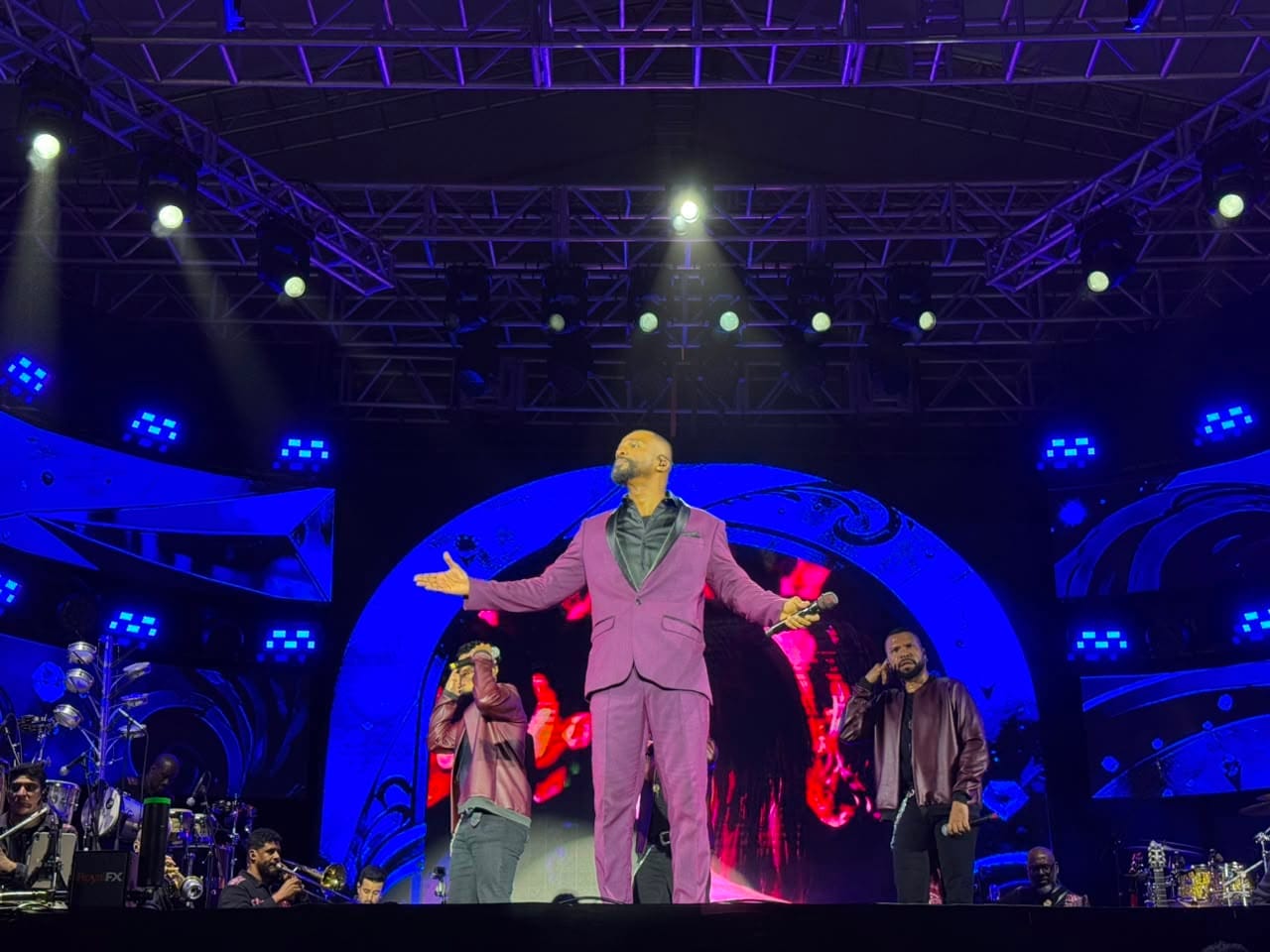 Close de Alexandre Pires no palco, dan&ccedil;ando e cantando com terno roxo e chap&eacute;u preto sob ilumina&ccedil;&atilde;o azul, com dan&ccedil;arinos fazendo coreografia ao lado.