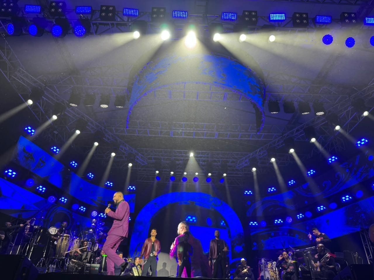 Close de Alexandre Pires no palco, cantando com terno roxo sob ilumina&ccedil;&atilde;o azul, com a banda e dan&ccedil;arinos desfocados ao fundo.