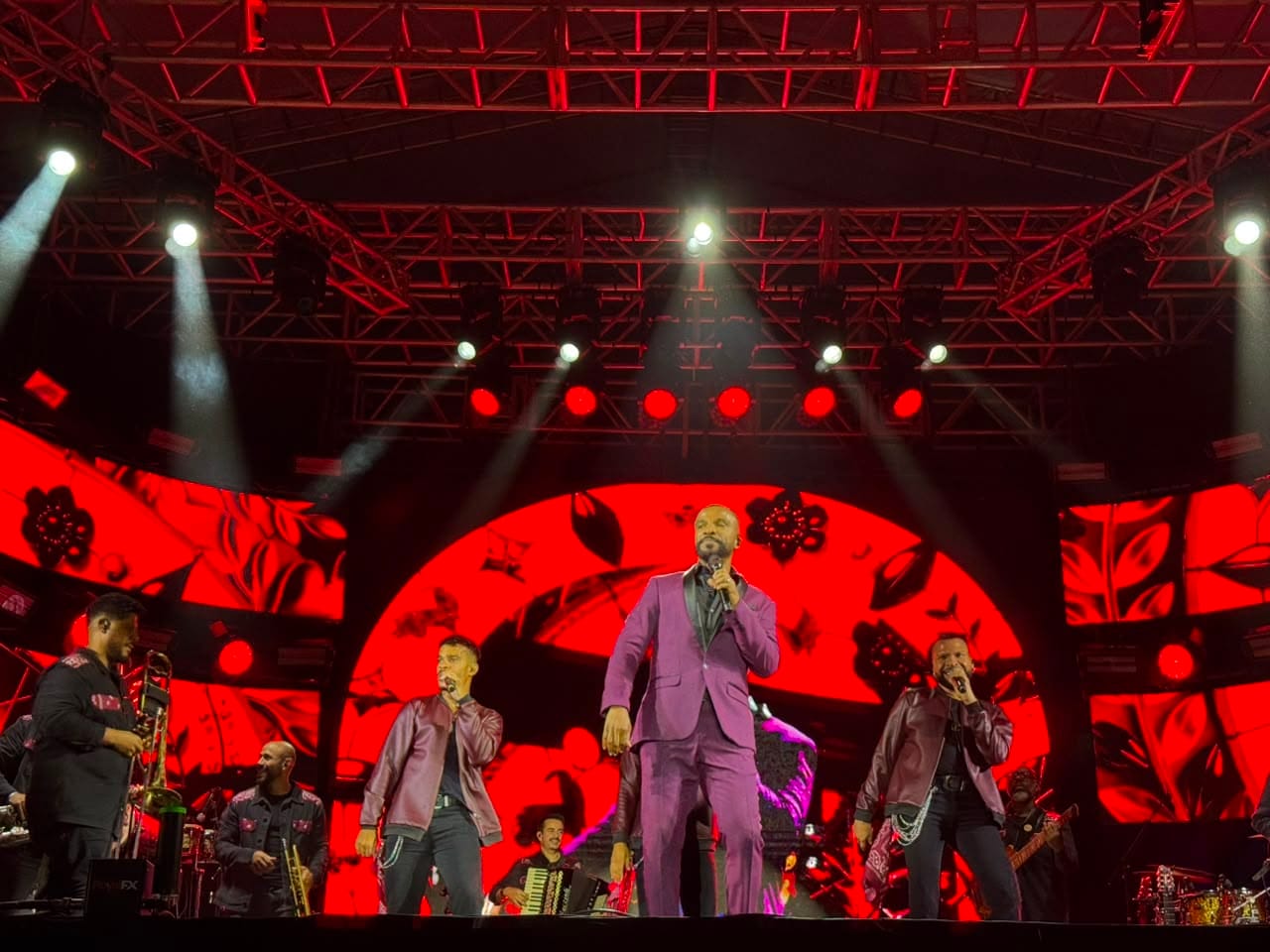 Alexandre Pires canta e dan&ccedil;a no centro de um palco iluminado por luzes azuis e verdes, ladeado por dois dan&ccedil;arinos. Ao fundo, um grande painel de LED exibe gr&aacute;ficos circulares.