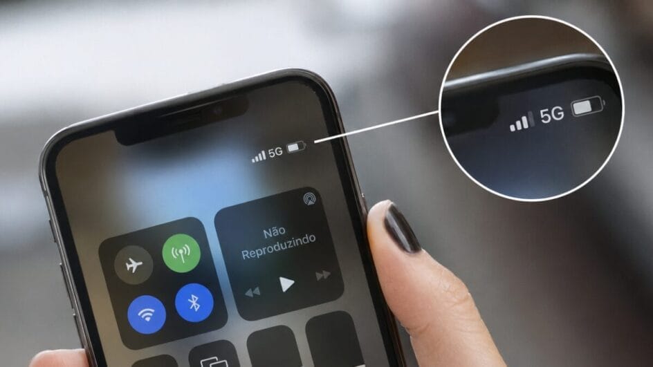 Close-up de uma pessoa segurando um smartphone com a Central de Controle aberta. No canto superior da tela aparece o indicador de conexão 5G ao lado das barras de sinal e do ícone de bateria. Um círculo ampliado destaca a área onde fica o símbolo da rede móvel, ilustrando as letras que identificam o tipo de conexão de internet no celular.