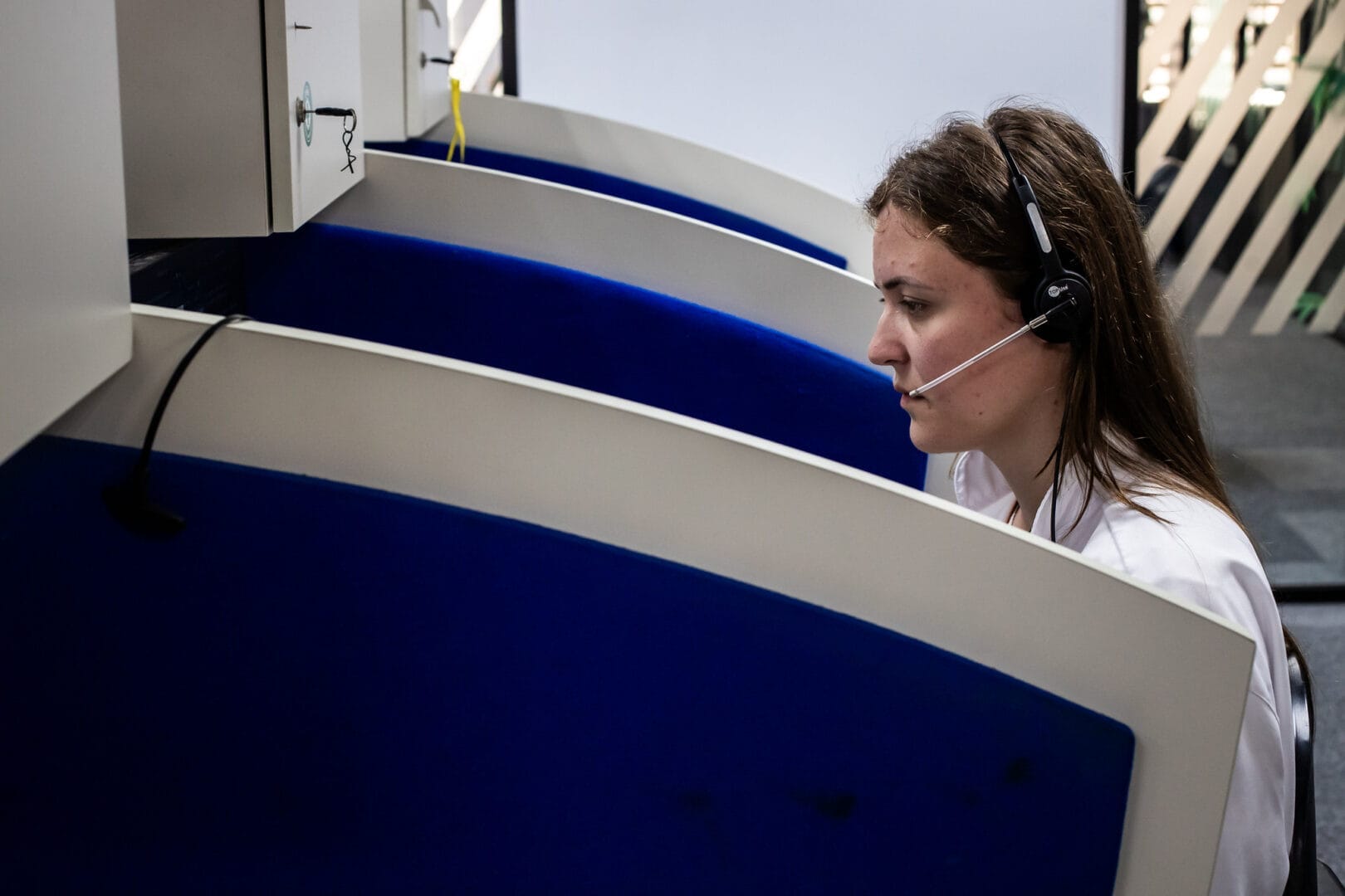 Mulher jovem com fone de ouvido e microfone acoplado trabalha sentada de perfil em uma cabine de atendimento de telemarketing.