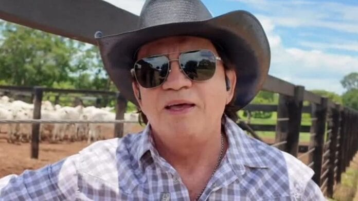 Chitãozinho de chapéu de cowboy e óculos escuros falando à câmera, com gado nelore ao fundo