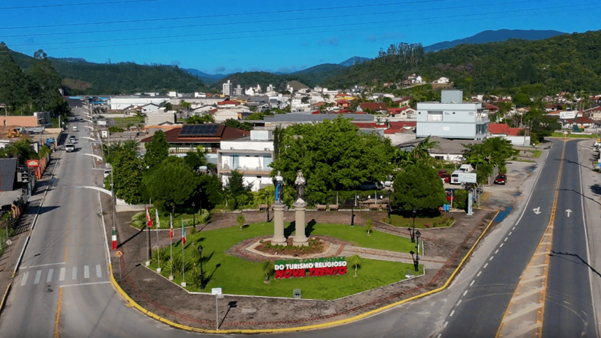 Vista aérea da área central de Nova Trento, em Santa Catarina, com praça, ruas, construções e morros cobertos pela vegetação ao fundo.