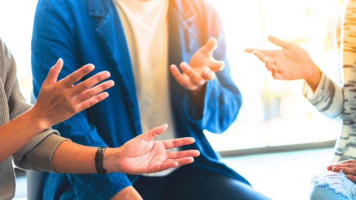 Fotografia de pessoa gesticulando durante conversa ou apresentação, em ambiente cotidiano, mãos visíveis e em movimento.