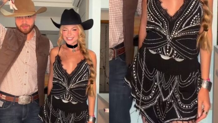 Mulher branca, loira, com chapéu preto, gargantilha preta, vestido preto com pedrarias e um cinto básico preto