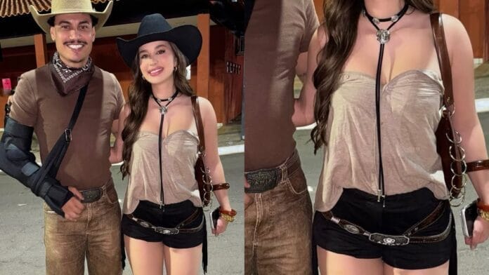 Mulher branca, com cabelo castanho, chapéu preto na cabeça, gargantilha longa preta, um top cropped bege, short preto e cinto 