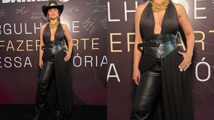 Mulher branca, de cabelo castanho, com chapéu preto, calça e top com decote na cor preta, corset preto e bota longa preta