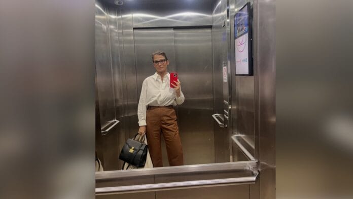 Jornalista Renata Vasconcellos, de cabelo castanho e óculos de armação escura, com blusa branca e calça marrom