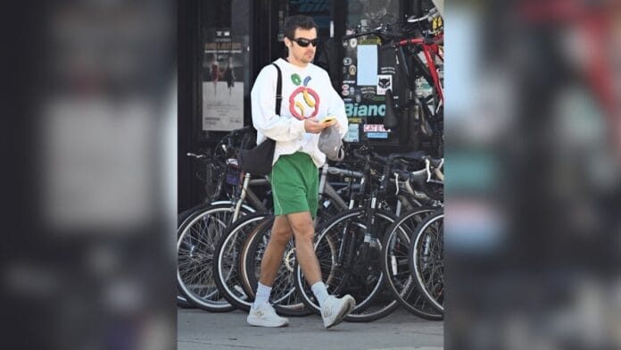 Homem com camiseta estampada, bermuda verde e tênis branco. Além de uma bolsa e óculos pretos