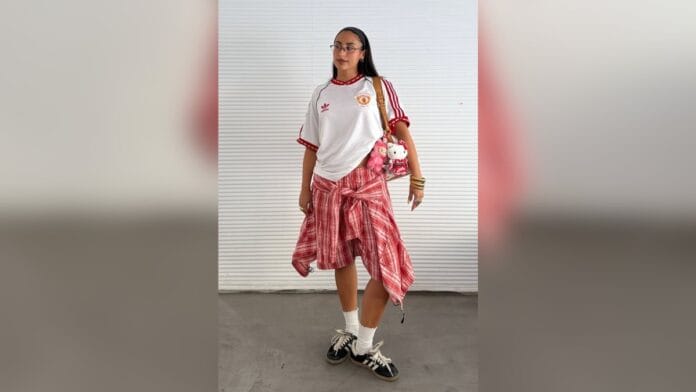 Mulher de cabelo castanho com blusa de time de futebol, camisa xadrez amarrada na cintura e tênis preto nos pés