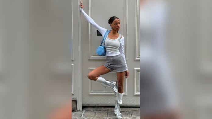 mulher de cabelos castanhos usando uma regata branca, casaco bolero branco, bolsa azul, short cinza e tênis prateado