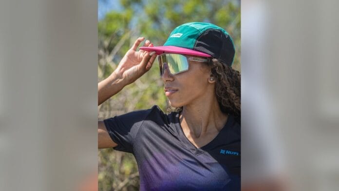 Mulher de cabelo castanho claro com boné verde, óculos espelhado e camiseta azul