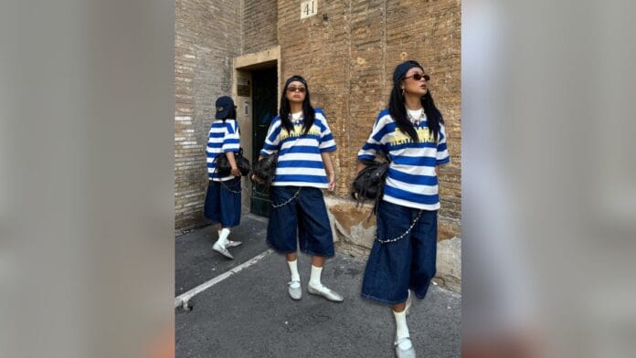 Mulher de cabelo solto preto, com camisa larga listrada e bermuda jeans comprida, sapatilha prateada nos pés 