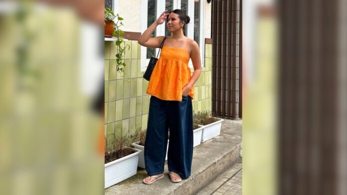 mulher com bata laranja, calça jeans wide leg e chinelo bege nos pés