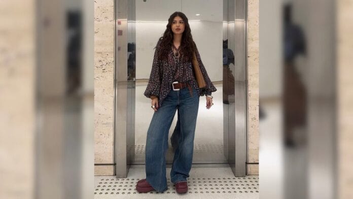 Mulher com bata estampada, cinto de couro marrom, calça jeans azul wide leg e bota roxa