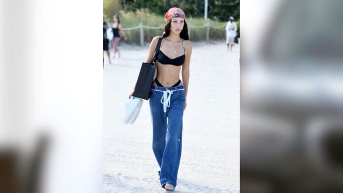 Mulher com calça cintura baixa jeans azul médio, com um cordão branco amarrado na cintura e biquini preto por baixo