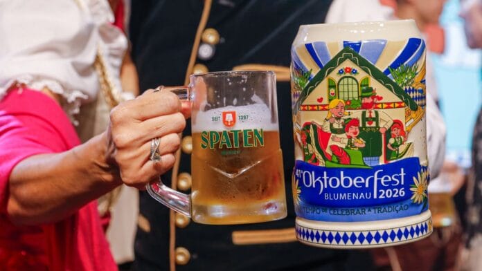 Mão segurando uma caneca de vidro com chope Spaten ao lado de uma caneca de cerâmica comemorativa da Oktoberfest Blumenau 2026, decorada com casal em trajes típicos alemães e casa enxaimel.