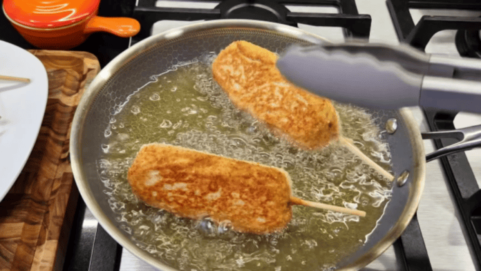 Dois espetinhos de hot dog coreano sendo fritos em óleo quente em uma frigideira