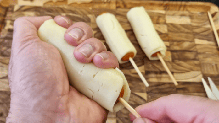 Mãos envolvendo uma salsicha no palito com uma fatia de queijo