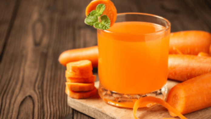 Copo com suco alaranjado de cenoura sobre mesa de madeira, com cenouras ao fundo
