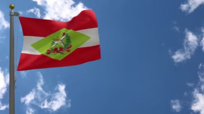 Bandeira do Estado de Santa Catarina hasteada num mastro sob um céu azul com nuvens, representativa da apuração geral das Eleições de 2026.