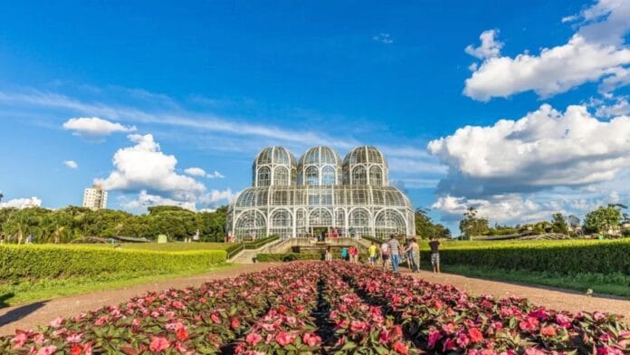 Estufa de vidro do Jardim Botânico de Curitiba rodeada por flores sob céu azul, representando a apuração para o Senado no Paraná nas Eleições 2026.