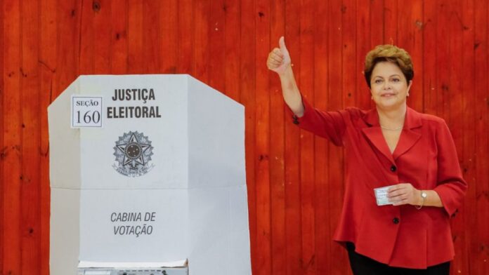Dilma Rousseff vestindo blazer vermelho faz sinal de positivo com o polegar ao lado de uma cabine de votação.