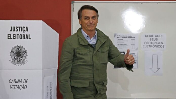 Jair Bolsonaro com jaqueta verde-oliva faz sinal de positivo ao lado da cabine de votação da Justiça Eleitoral.