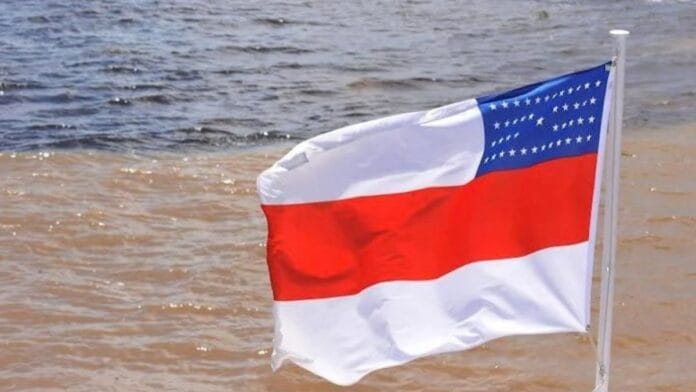 Bandeira do estado do Amazonas com suas cores branca, vermelha e azul com estrelas, hasteada e ondulando com o rio ao fundo, representando a apuração das Eleições 2026