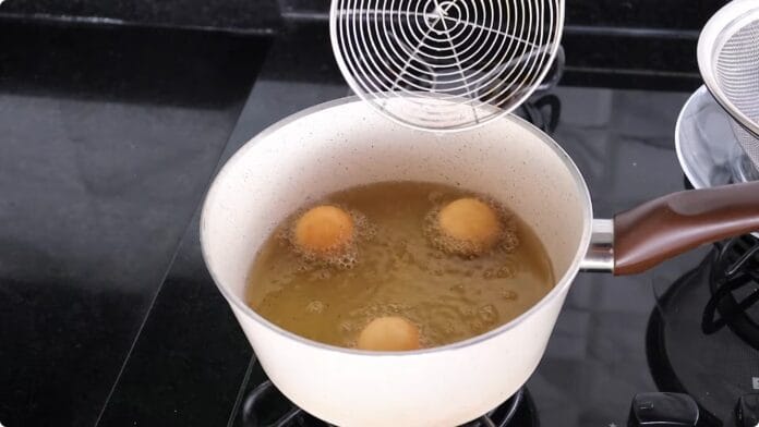 Três bolinhos de chuva dourados fritando em óleo, com escumadeira acima da panela