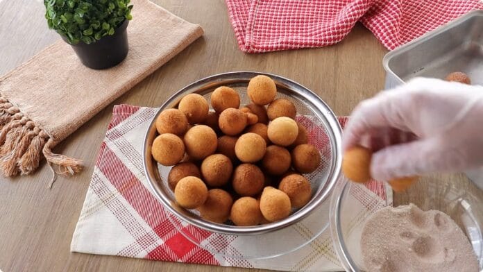 Mão pegando bolinho dourado ao lado de recipiente com açúcar e canela