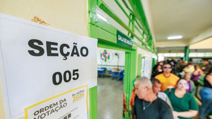 Cartaz com a inscrição Seção 005 na porta de uma sala com eleitores organizados em fila no corredor para votar.