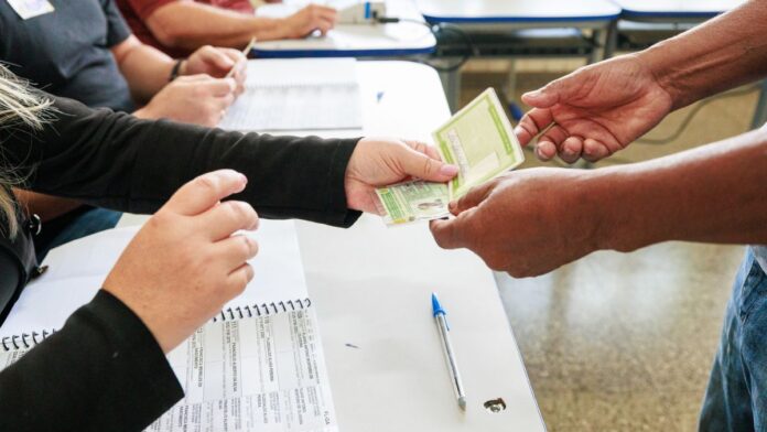 Mãos de um eleitor entregando o título de eleitor em papel para a mesária sobre o caderno de votação em seção eleitoral.